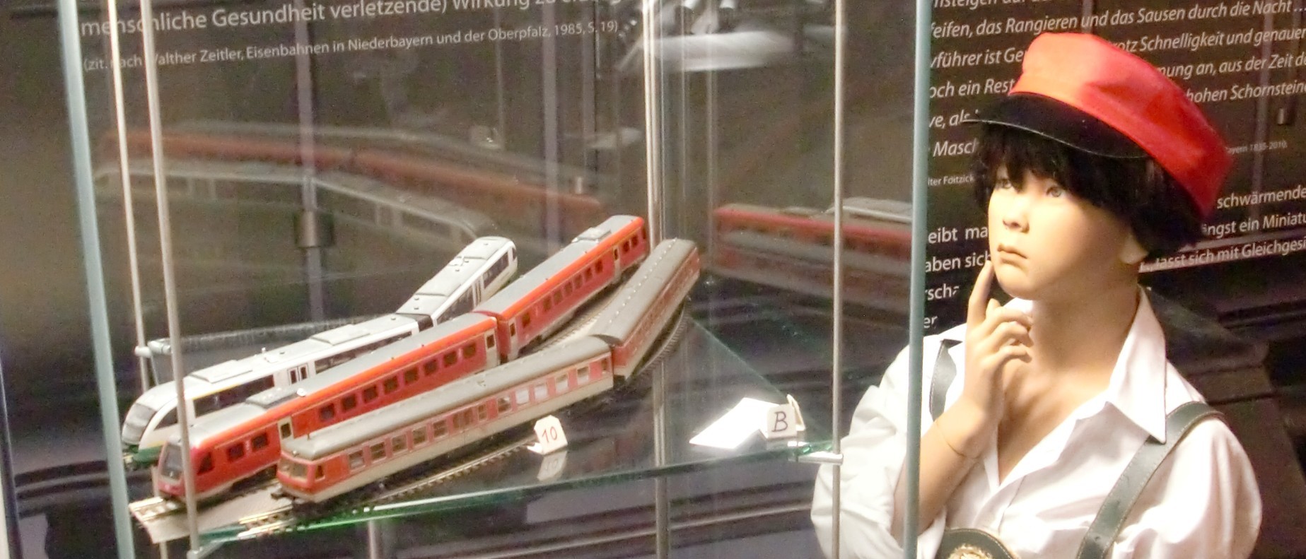 Das Foto zeigt einen Ausschnitt der Ausstellung &quot;150 Jahre Eisenbahn&quot;. Es ist eine kindliche Puppe mit Schaffnermütze zu sehen, die eine Vitrine mit Modelleisenbahnzügen betrachtet.