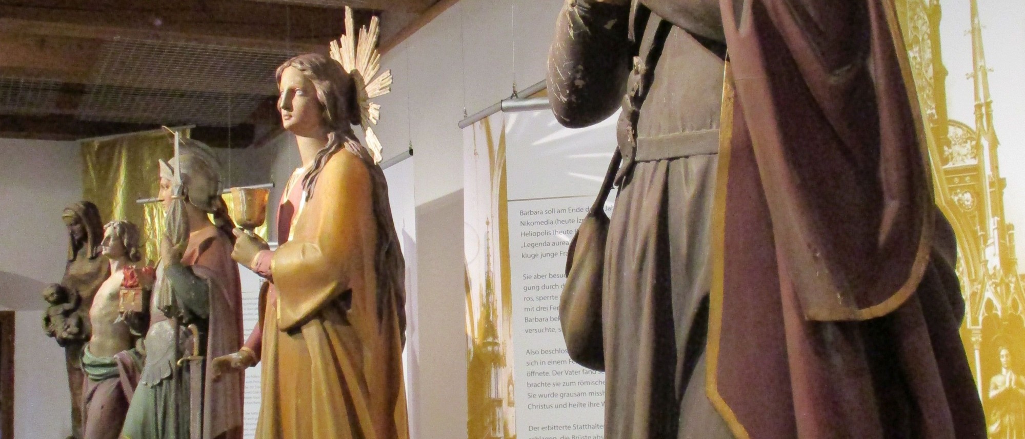 Das Bild zeigt einen Ausschnitt der Ausstellung &quot;Aller Heiligen&quot;. Es sind lebensgroße Figuren der hll. Barbara, Florian und Sebastian zu sehen. Im Hintergrund eine Maria mit Kind.