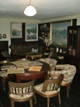 Das Foto zeigt einen Ausschnitt des Wohnzimmers von Karl Schmidt. Man sieht Bilder und andere Kunstobjekte an den Wänden sowie ein Sofa, Regale, Stühle und einen Mosaiktisch.